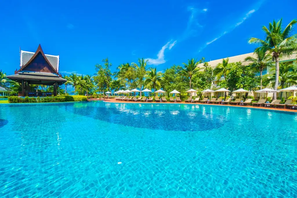 Large turquoise resort swimming pool with sun loungers, palm trees, and a Thai pavilion under a clear blue sky.