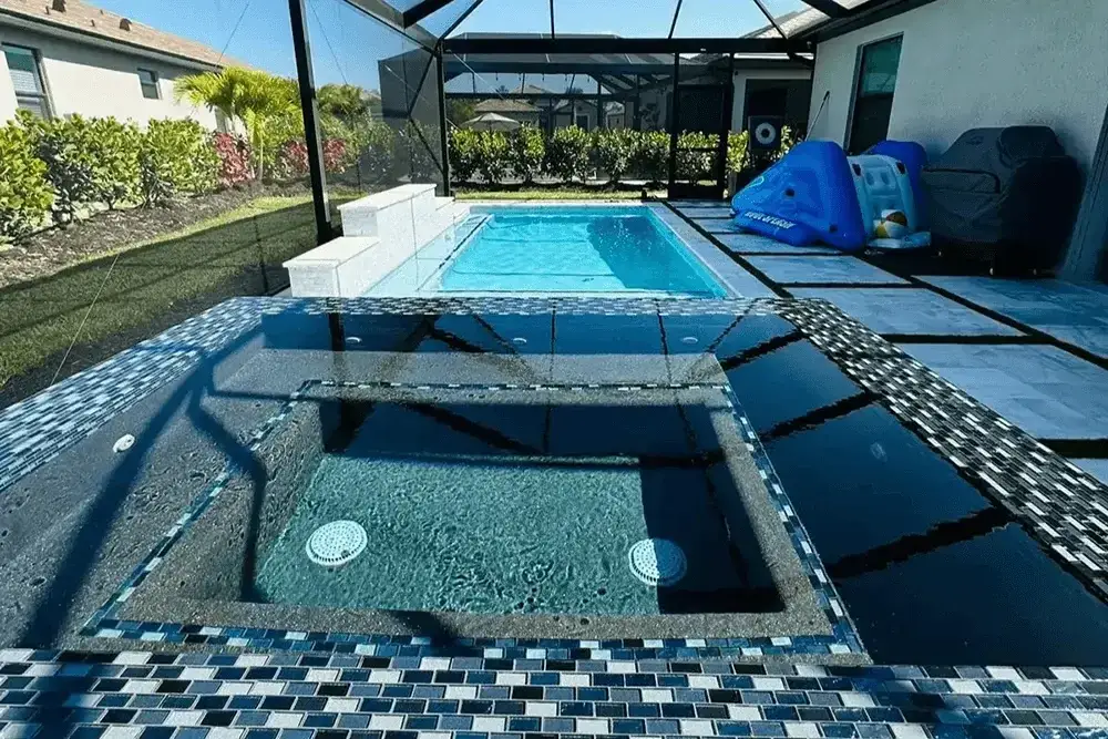 Inground pool with mosaic-tiled spa and blue water toys on patio, surrounded by landscaping and a screened enclosure.
